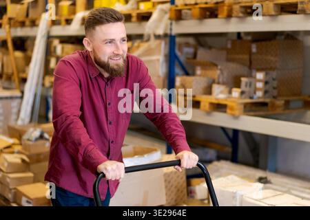 Portrait of a happy male logistics manager in a red shirt operating a manual trolley. Professional warehouse worker smiling while handling inventory delivery in a storage facility. || Model released Stock Photo