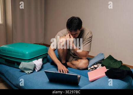 Young backpacker male sitting on a sofa, experiencing stress while packing a suitcase and using a laptop, representing the challenges of planning a Stock Photo