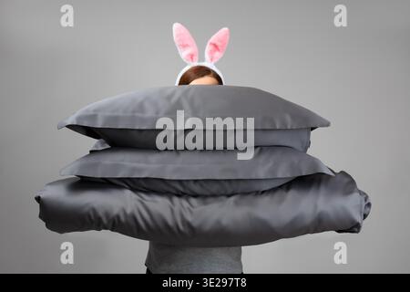 A woman in Easter bunny ears holds a warm duvet and gray feather pillows. Bed linen for sleeping. || Model released Stock Photo