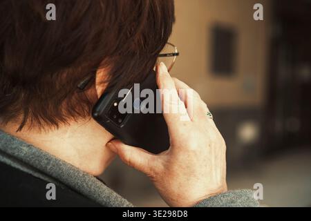 Senior woman talking on smartphone outdoors, back view with copy space. Active elderly lifestyle and mobile communication concept. Stock Photo