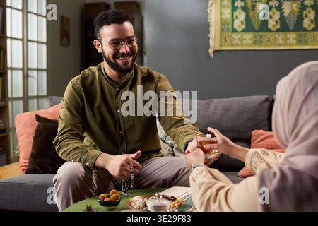 Young adult Middle Eastern man smiling while sitting on sofa holding prayer beads, reaching for glass offered by Middle Eastern woman in hijab, sharing traditional snacks during gathering Stock Photo