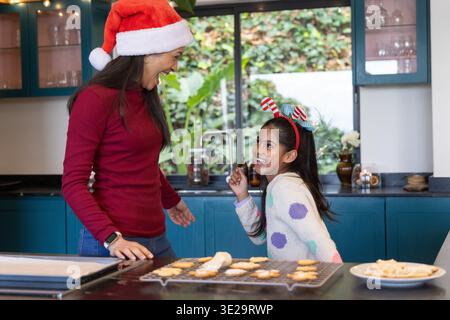 Asian mother and child baking cookies on kitchen countertop with cooling rack, dough log, Santa hat. Family, festive, home, cozy, baked, biscuits, jar Stock Photo