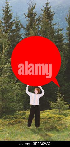 Man standing in forest landscape holding oversized red speech bubble shape above head. Stock Photo