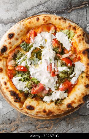 Homemade pizza with stracciatella cheese, pesto, ham and cherry tomatoes close-up on a wooden board on the table. Vertical top view from above Stock Photo