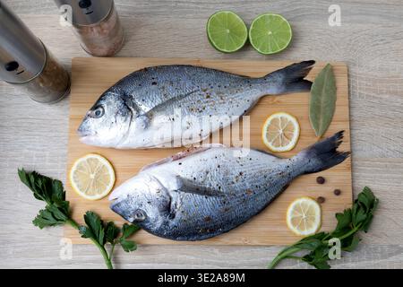 Two Fresh Fish on a Cutting Board with Spices and Herbs. Two whole, fresh fish are laid out on a wooden cutting board, surrounded by slices of lemon Stock Photo