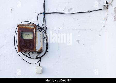 An old-style electric meter in a rusty case on a white wall with switches and wires Stock Photo