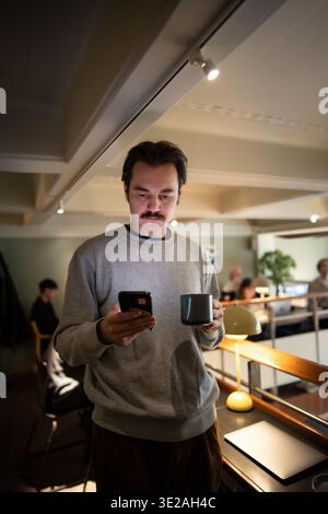 Handsome businessman enjoying coffee and scrolls smartphone after finishing work at co-working cafe. Stock Photo