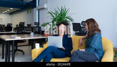 Two women discussing work on a yellow sofa in a contemporary office setting Stock Photo