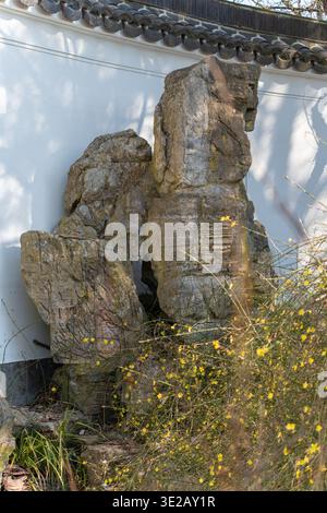 White wall with dark-tiled roofline. Three mossy rocks lean against the wall. Tiny carved inscription appears on one stone. Yellow wildflowers grow at Stock Photo