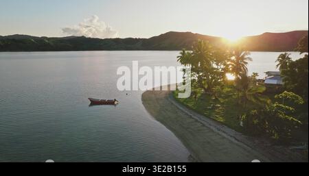 Small red boat anchored near a sandy beach with palm trees and houses, capturing the warm glow of the sun setting over calm ocean waters and distant hills in Fiji Stock Photo