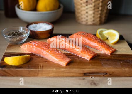 salmon fillets seasoned with sea salt crystals and cracked black pepper, arranged on a two-tone wooden cutting board with lemon wedges. Stock Photo