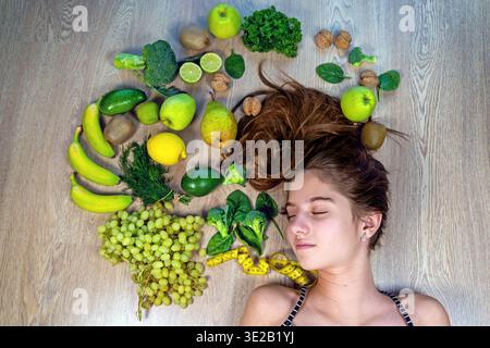 Young woman surrounded by healthy fruits. A serene young woman with closed eyes lying on a wooden floor, surrounded by fresh green fruits, vegetables Stock Photo