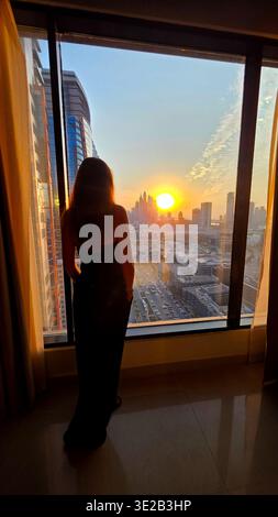 Woman watching Dubai sunset from hotel window. Silhouette of a woman standing by a large hotel window, overlooking Dubai skyline at sunset with warm Stock Photo