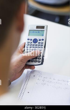 Student holds a scientific calculator and calculates on a task, next to it are notes with mathematical formulas in a checkered notebook Stock Photo