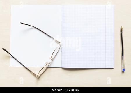 Top view of pen and eyeglasses on blank open school notebook with squared sheets with margins on light brown wooden board Stock Photo