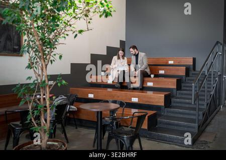 Partners going over notes together on wooden office steps in plant-filled workspace. Notes, plant-filled workspace, office, teamwork, partners. Stock Photo