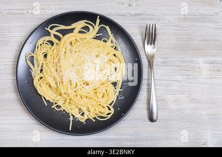 Italian cuisine - top view of spaghetti al burro e parmigiano (pasta with butter and cheese) on black plate and fork on gray wooden background with bl Stock Photo