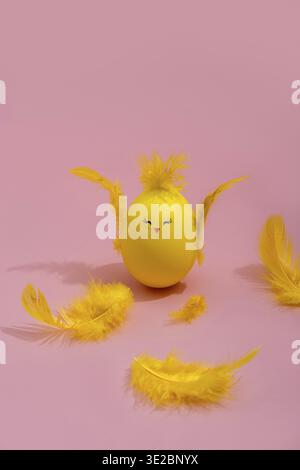 Cute yellow Easter egg in the shape of chick surrounded by feathers on a bright pink background. Easter greeting card Stock Photo