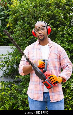 African American man holding hedge trimmer while wearing goggles, earmuffs, orange gloves in yard. Gardening, landscaping, outdoors, cordless, pruning Stock Photo