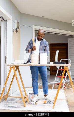African American man stirring paint on plank workbench at front porch with two buckets, denim shirt. Painter, outdoor, renovation, ladder, entryway, t Stock Photo