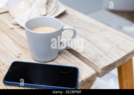 Displaying light gray ceramic mug sitting on home slatted side table, with blue-cased smartphone. Woodgrain, rustic, latte, device, gadget, calm, mini Stock Photo