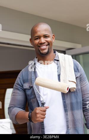 African American man standing in doorway holding paint roller, wearing white tee, towel on shoulder. Painter, homeowner, renovation, entryway, natural Stock Photo