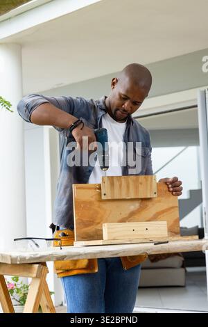 African American man drilling and assembling wood frame on front porch using cordless drill. Handyman, woodworking, powertool, workbench, outdoor, day Stock Photo