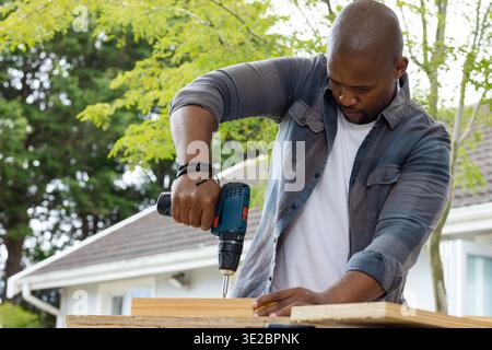 African male using cordless drill on wooden plank on sawhorse in house backyard, wearing gray shirt. Outdoor, yard, woodworking, handyman, timber, boa Stock Photo