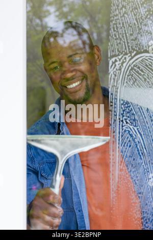 African man cleaning large glass window at home with hand squeegee, smiling in denim jacket. Pane, reflections, greenery, closeup, minimalist, domesti Stock Photo