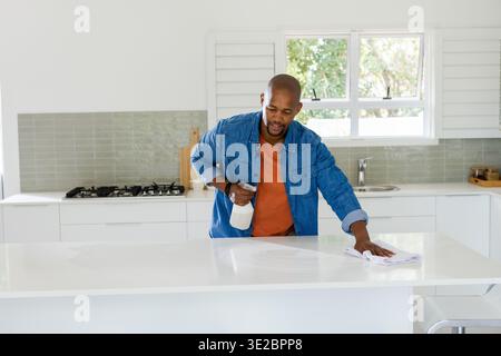 African American man cleaning white kitchen island with spray bottle and cloth wearing denim jacket. Countertop, wipe, modern, bright, natural, greene Stock Photo