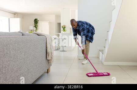Man cleaning tiled floor with pink flat mop in modern living room near sofa, copy space. Tidy, household, interior, minimalist, airy, neutral, dayligh Stock Photo