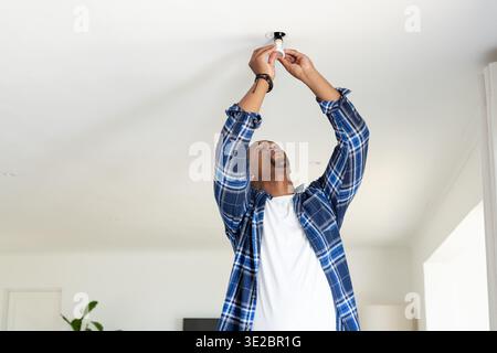 African American man wearing blue plaid shirt changing ceiling light bulb in living room. Home, interior, renovation, maintenance, installation, fixtu Stock Photo