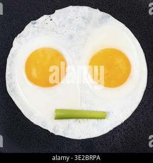 Fried eggs and piece of celery on black plate close up. Fried eggs like face with straigh closed mouth Stock Photo