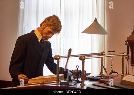 A lifelike wax figure of a bearded Victorian businessman in a dark suit stands at an antique wooden desk, examining a large open ledger. The... Stock Photo