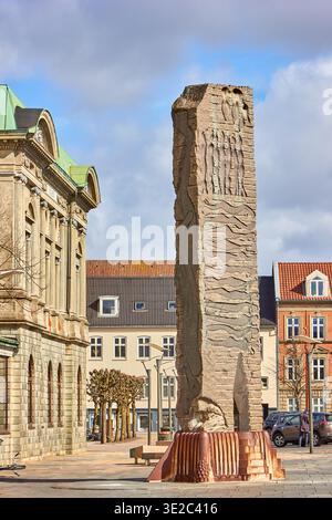 A tall rectangular stone monument featuring intricate carved reliefs stands prominently in a historic European town square. The monument, mounted... Stock Photo