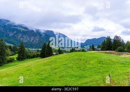 A scenic alpine valley with rolling green meadows, scattered trees, and small rural houses surrounded by mountains. Low clouds and mist drift across t Stock Photo