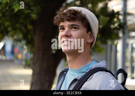 Late-teen male standing on city sidewalk near large tree, wearing knit beanie and carrying backpack. Urban, outdoors, daylight, student, hat, bag, cas Stock Photo