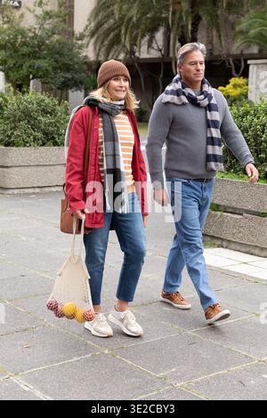 Middle-aged couple walking on plaza with scarves, carrying mesh bag with fruit and shoulder bag. Urban, duo, casualwear, knitwear, overcast, cool, gro Stock Photo