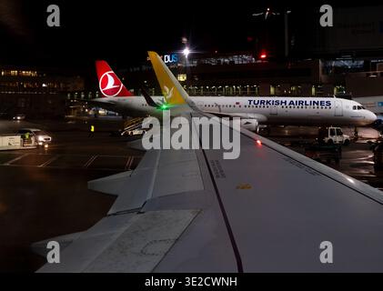 View from a passenger window over the wing to a Turkish Airlines aircraft on a wet night apron with ground service vehicles and lit terminal at Düssel Stock Photo