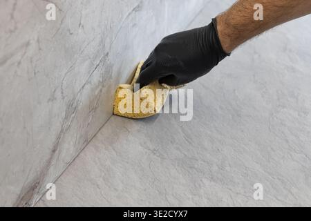 Worker in a black nitrile glove cleaning grout residue from gray porcelain tiles with yellow sponge during kitchen renovation. Construction finishing Stock Photo