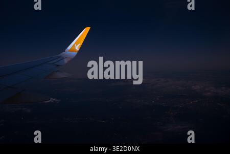Commercial airplane wing gliding over glowing city lights under a dark night sky and scattered clouds viewed from a passenger window in flight. Stock Photo