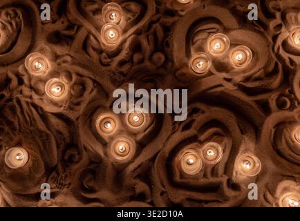 Top view of small candle lights glowing in sculpted brown sand texture creating an abstract warm background with soft shadows cozy ambience and decora Stock Photo