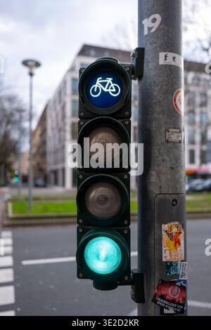 Close-up of a bicycle traffic light glowing green on an urban street with a sticker covered pole and soft focus buildings in the background. Stock Photo