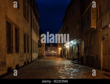Quiet narrow cobblestone street between historic buildings at night with warm window light and a deep blue sky creating a moody atmospheric Stock Photo
