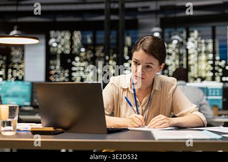 Female accountant in office using laptop, writing down information, working overtime to finish paperwork before deadline. Woman transcribing from device, doing note taking, documenting research Stock Photo
