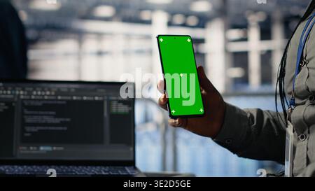Engineer using isolated screen smartphone in server room while optimizing deep learning systems handling tasks. Data center worker uses chroma key mobile phone, adjusting AI model parameters, camera B Stock Photo