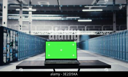 Green screen laptop in high tech data center facility with multiple rows of fully operational server racks. Mockup notebook used to oversee server hub built for digital storage systems Stock Photo