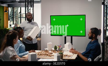 Property engineers team study architectural blueprints with a mockup on TV, collaborating in urban development master plan. Modern residential project with infrastructure goals. Stock Photo