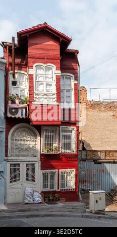 Vertical view of a narrow red wooden townhouse with white arched windows decorative iron gates potted plants and weathered urban surroundings under a Stock Photo