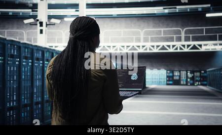 Engineer in server hub using laptop to upgrade rigs for increase in workload capacity using AI deep learning. Data center employee uses artificial intelligence to improve processing speed, camera A Stock Photo